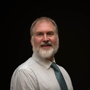 A white bearded middle aged man with a smiles at the camera, wearing a light shirt and patterned tie against a black ark background.