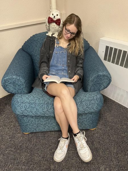 Catherine Brogan, a young woman with long, blonde hair and glasses, sits in a cozy teal chair with a leaf pattern. She is wearing a dark jacket over denim overalls and white sneakers. She is focused on reading a book on her lap, while a plush owl with a red bowtie is perched on her shoulder.