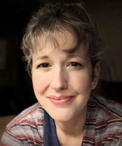 A white woman with short, wavy hair, smiling at the camera, wearing a striped shirt against a black background.